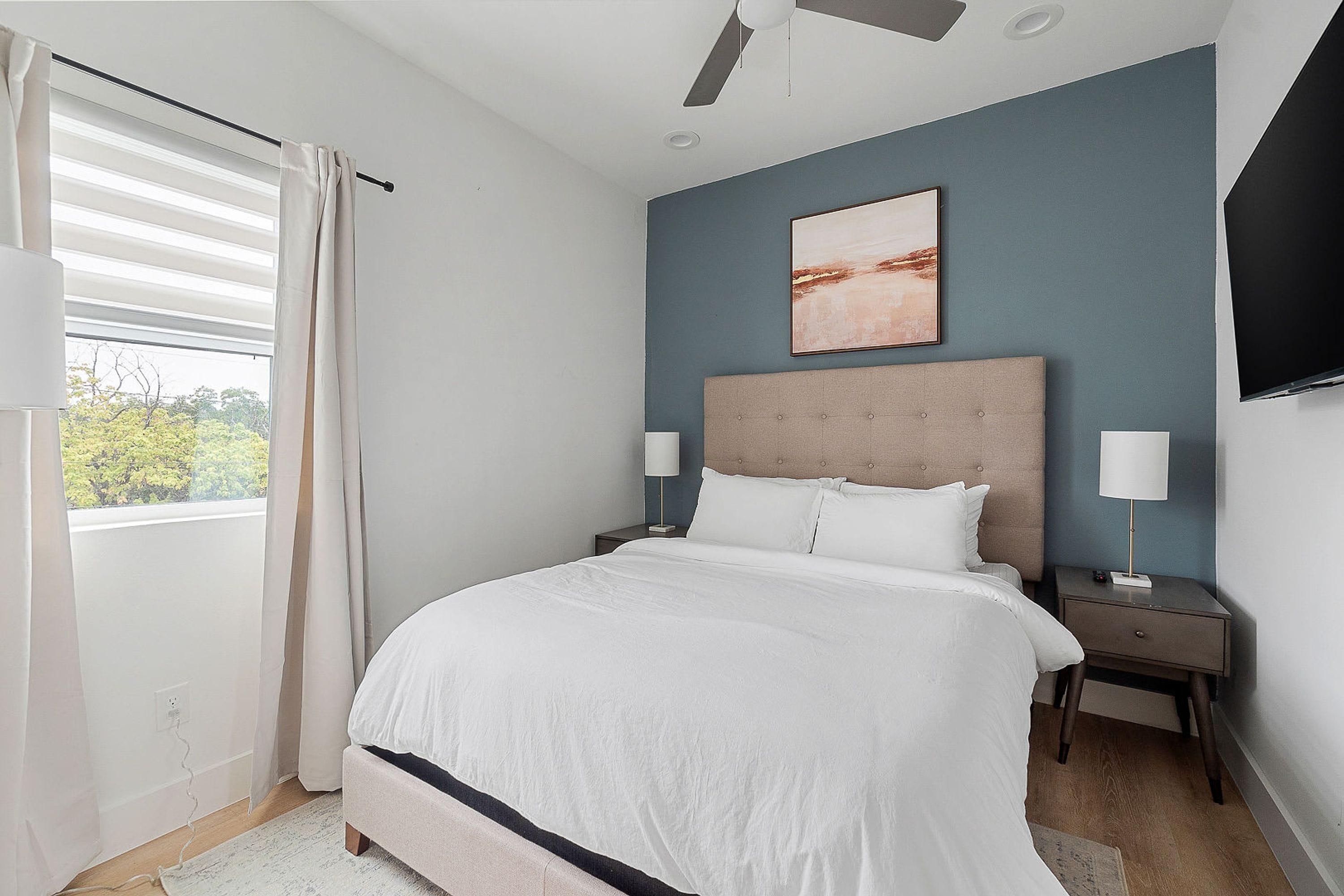 A bedroom with a bed, nightstands, a ceiling fan, a window with curtains, and a wall-mounted TV.