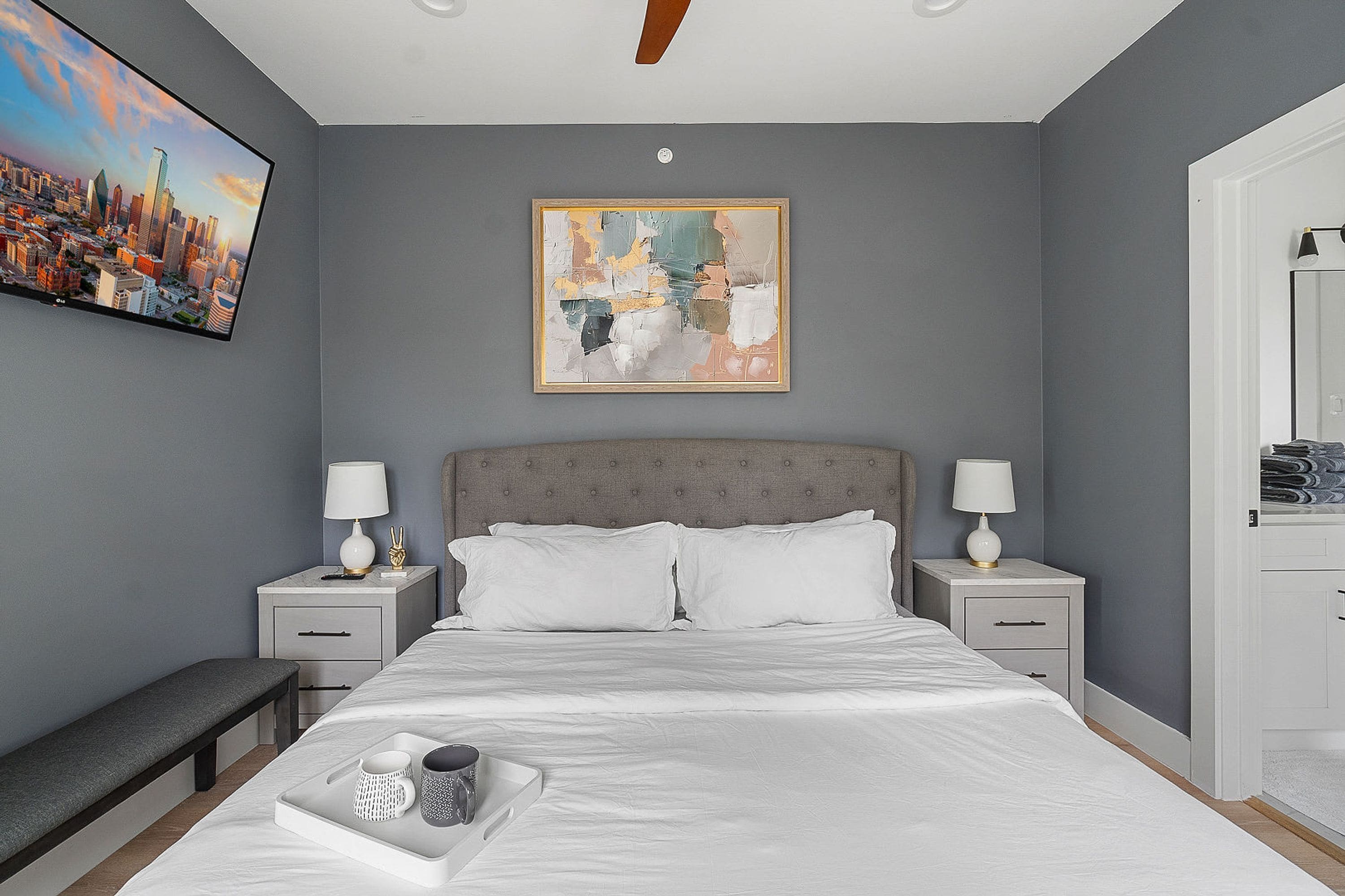 Une chambre à coucher avec un lit, deux tables de chevet avec des lampes, un banc, un plateau avec des tasses et un tableau représentant un paysage urbain accroché au mur.