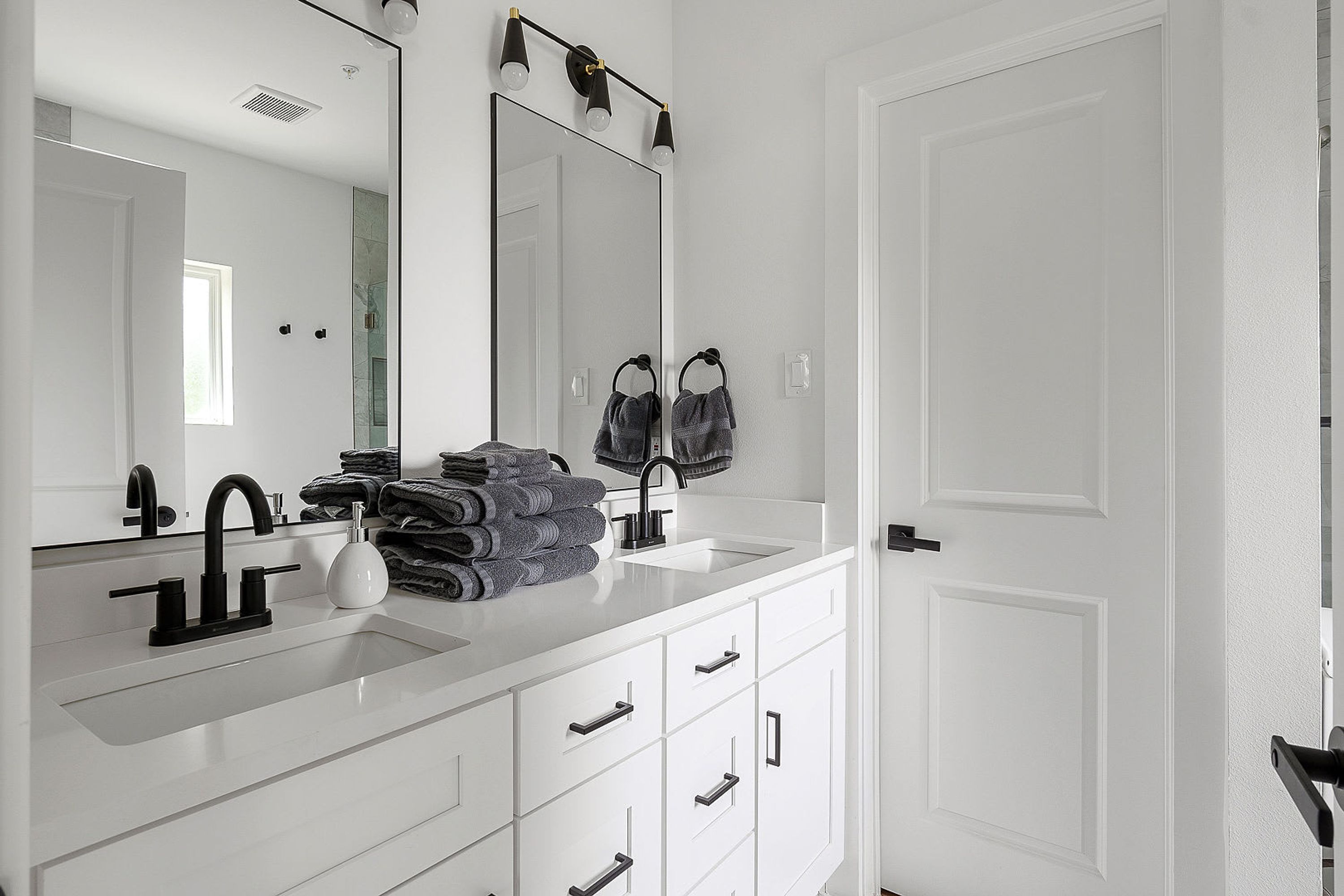 Une salle de bain moderne avec un meuble-lavabo blanc, un grand miroir et un évier. Des serviettes sont pliées sur le meuble-lavabo, et une porte se trouve sur la droite.