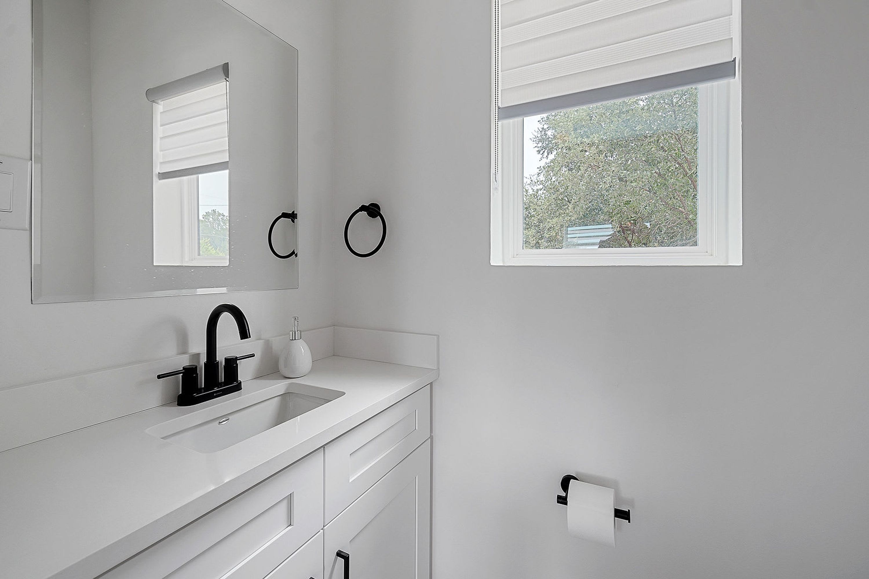 A modern bathroom with a white sink, black faucet, and a mirror above it. There are two towel rings on the wall and a window with blinds.