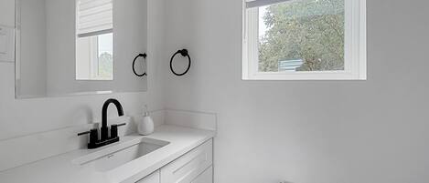 A modern bathroom with a white sink, black faucet, and a mirror above it. There are two towel rings on the wall and a window with blinds.