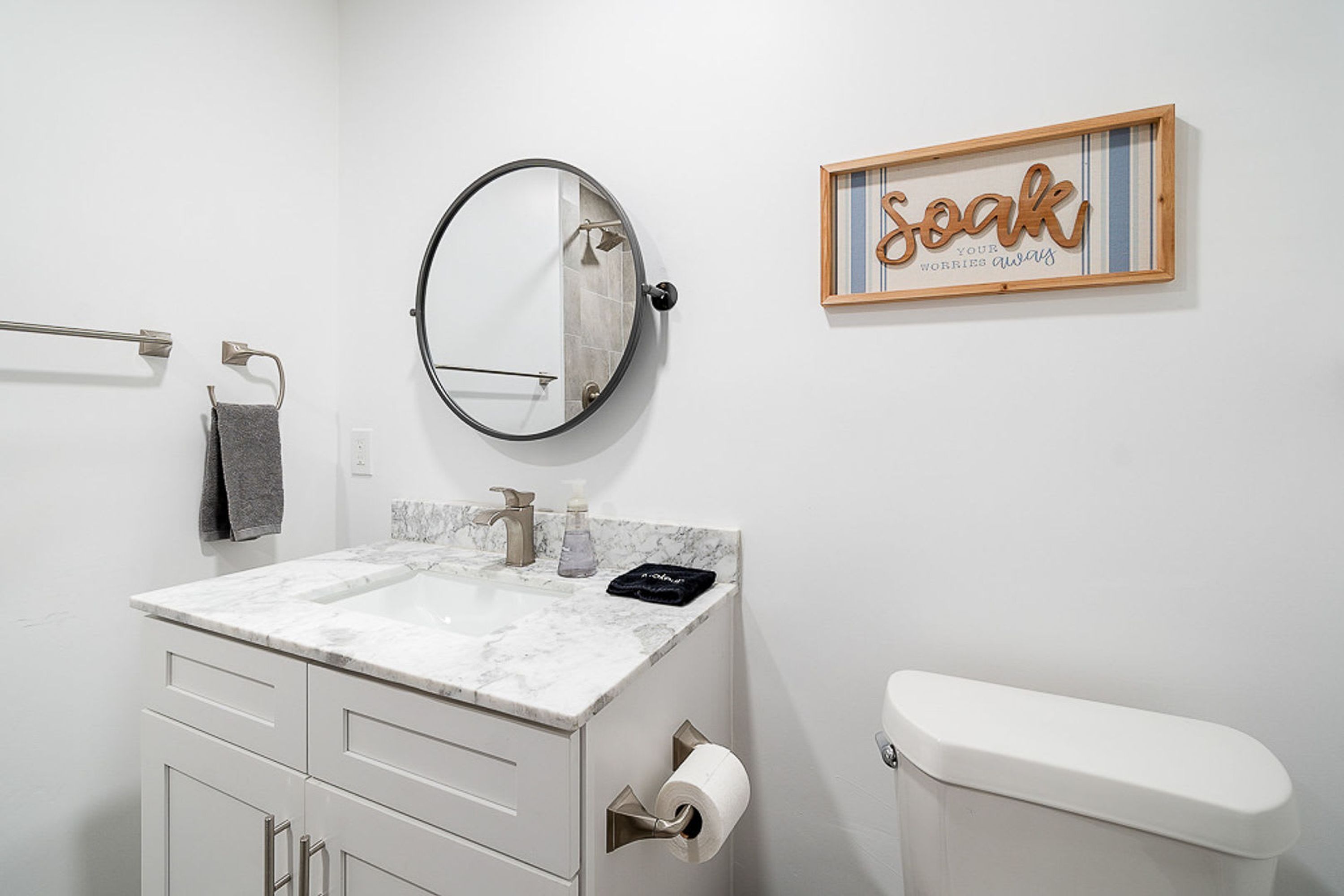 A bathroom with a white toilet, a sink with a marble countertop, a round mirror, a towel rack, and a framed sign on the wall.