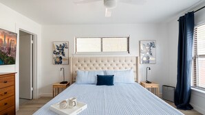 A bedroom with a large bed, a ceiling fan, a window with blinds, and artwork on the walls.