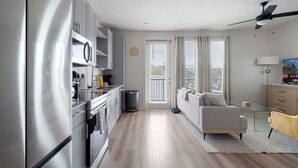 Una cocina moderna con electrodomésticos de acero inoxidable, una zona de comedor con un sofá y una mesita, y un balcón con cortinas.