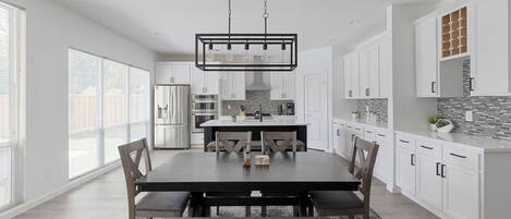 Une cuisine moderne avec des meubles blancs, un îlot central et un coin repas comprenant une table et des chaises aux couleurs sombres.