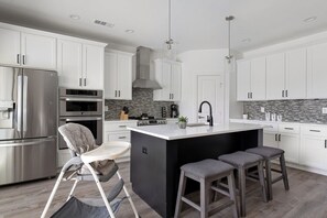 A modern kitchen with white cabinets, stainless steel appliances, a central island with stools, and a baby high chair. - Urban Retreat With Pool Near Downtown Dallas (Dallas)