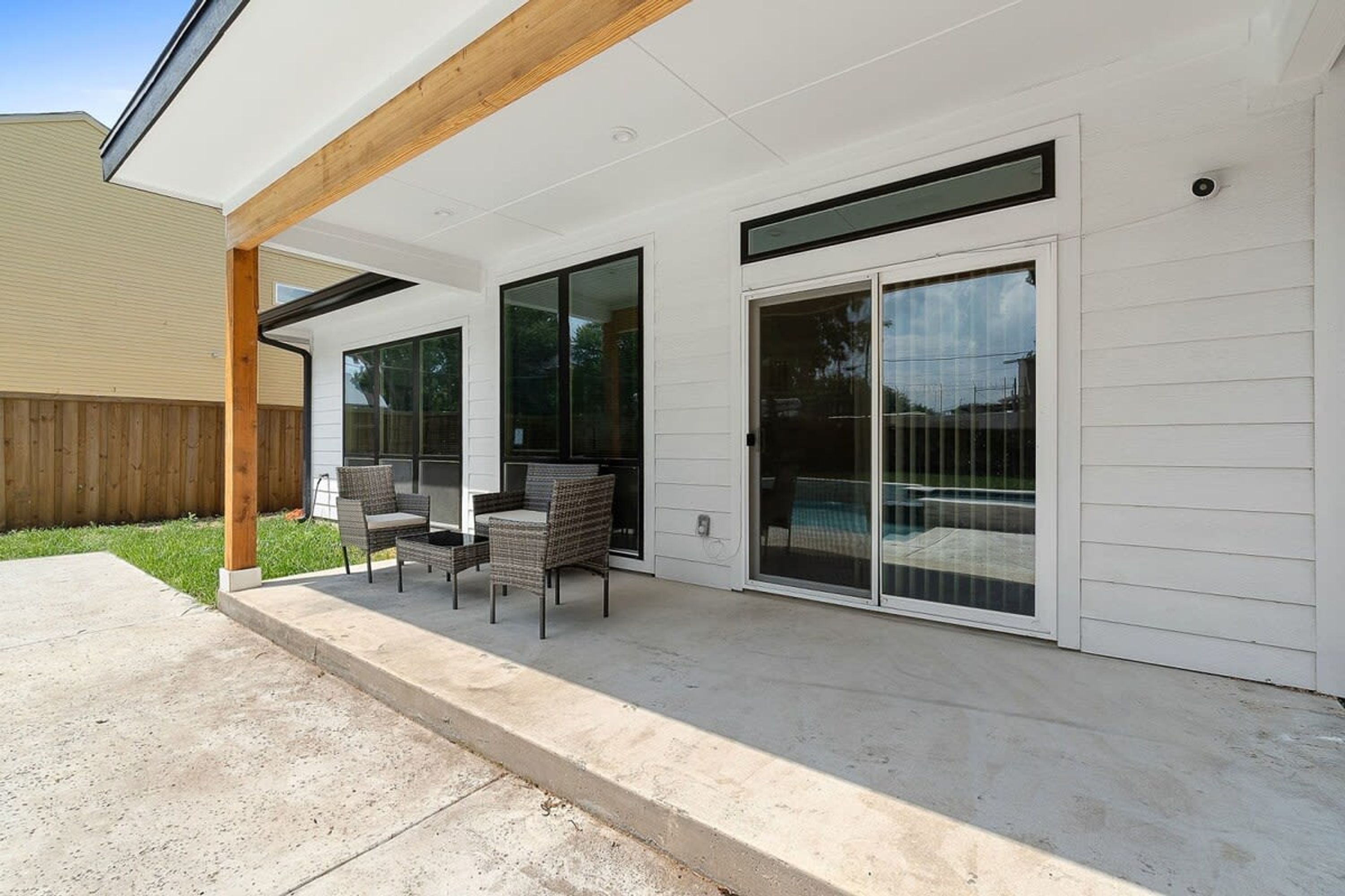 A covered patio with two wicker chairs and a table, large sliding glass doors, and a wooden beam ceiling.