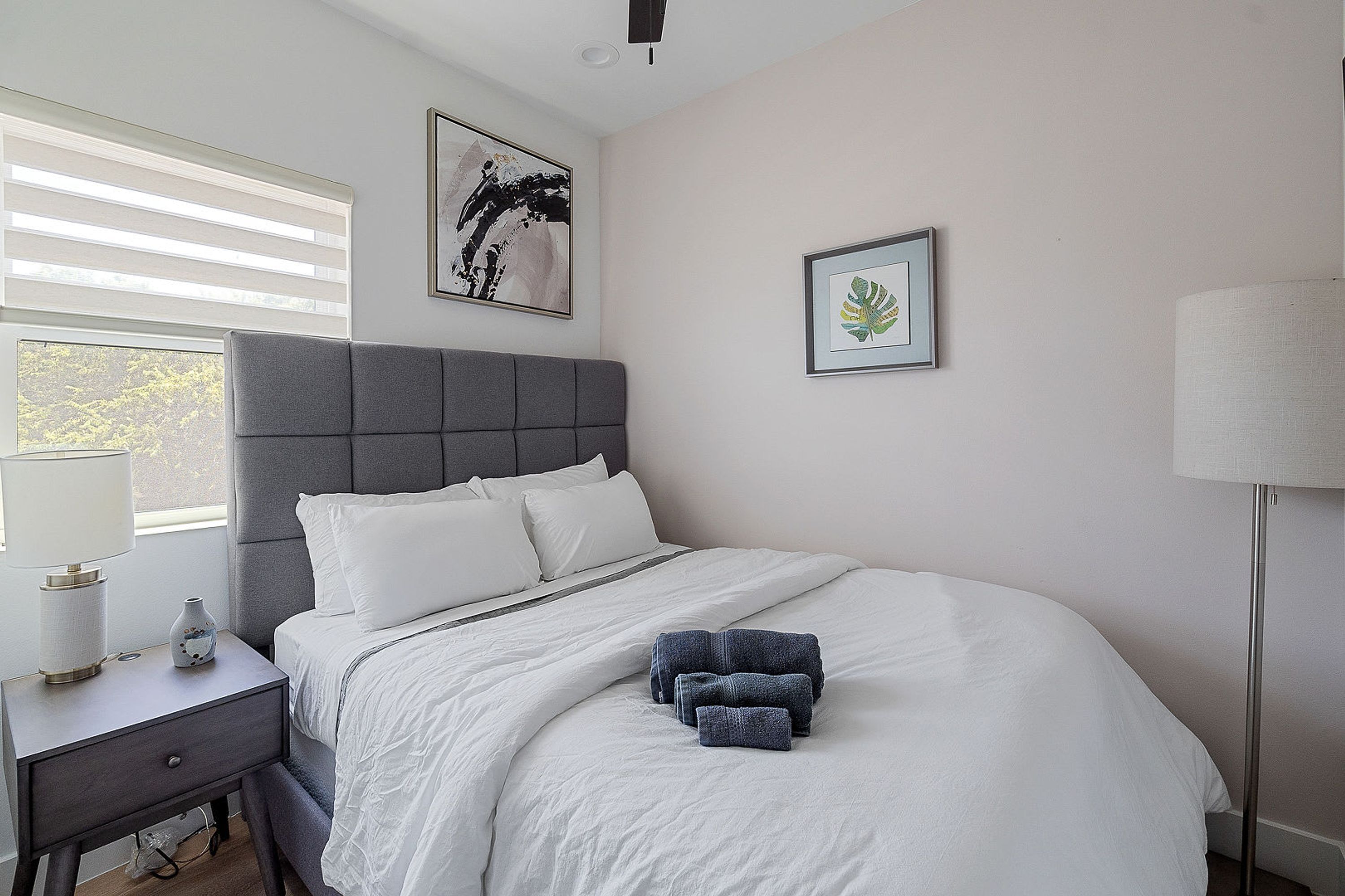 Un dormitorio con cama, mesita de noche, lámpara y una obra de arte en la pared.
