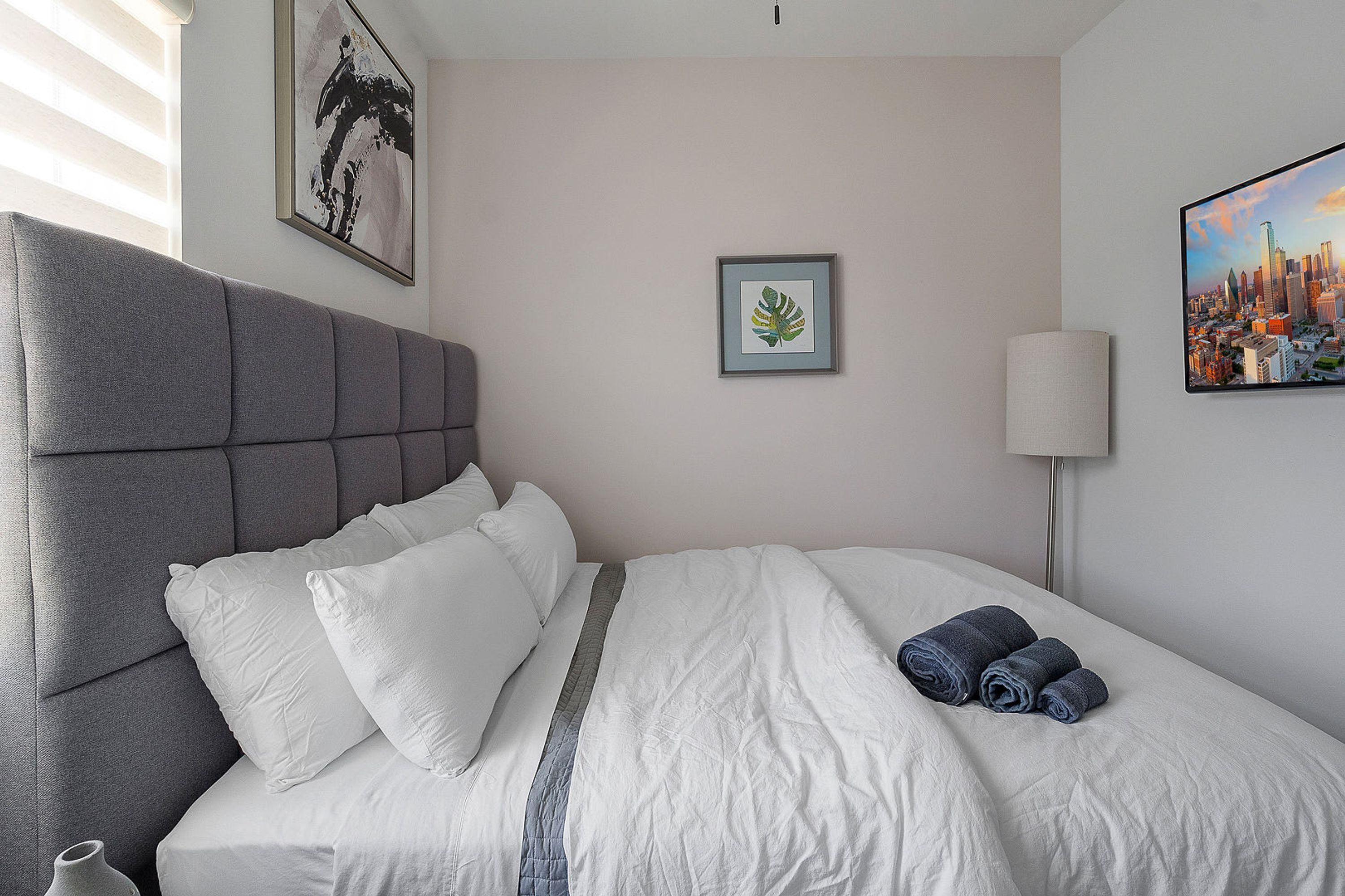 Una cama bien hecha con un cabecero gris, ropa de cama blanca y un cuadro de un paisaje urbano en la pared.