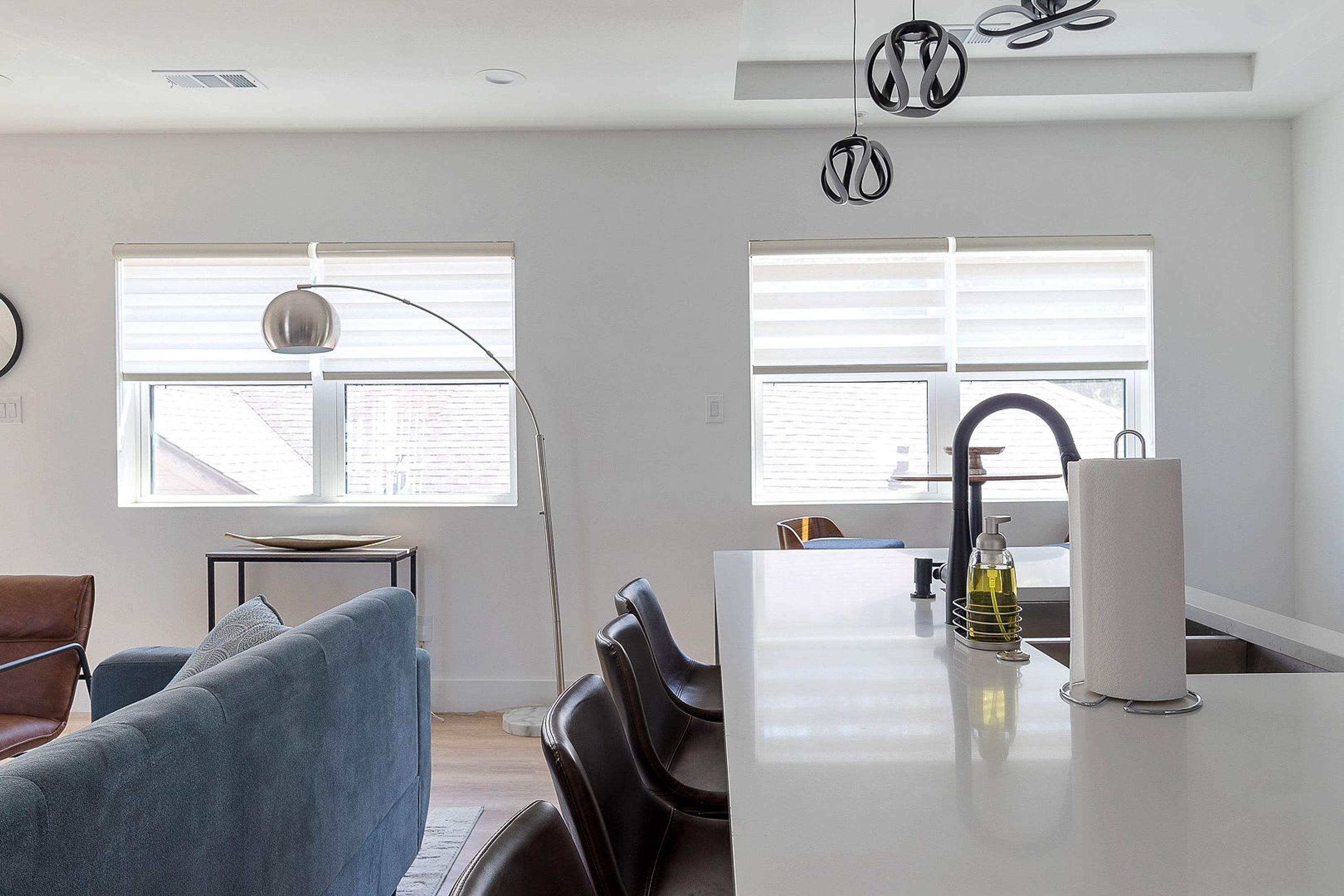 Eine moderne Küche mit weißer Arbeitsplatte, Spüle und Fenster mit Jalousien. Im angrenzenden Wohnbereich steht ein Stuhl und ein Tisch mit einer Lampe.