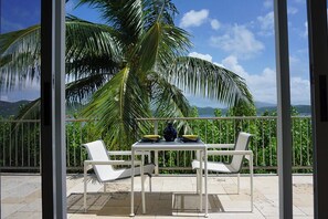 Ein Balkon mit zwei weißen Stühlen und einem Tisch, der auf eine tropische Landschaft mit Palmen und Blick auf das Meer blickt.