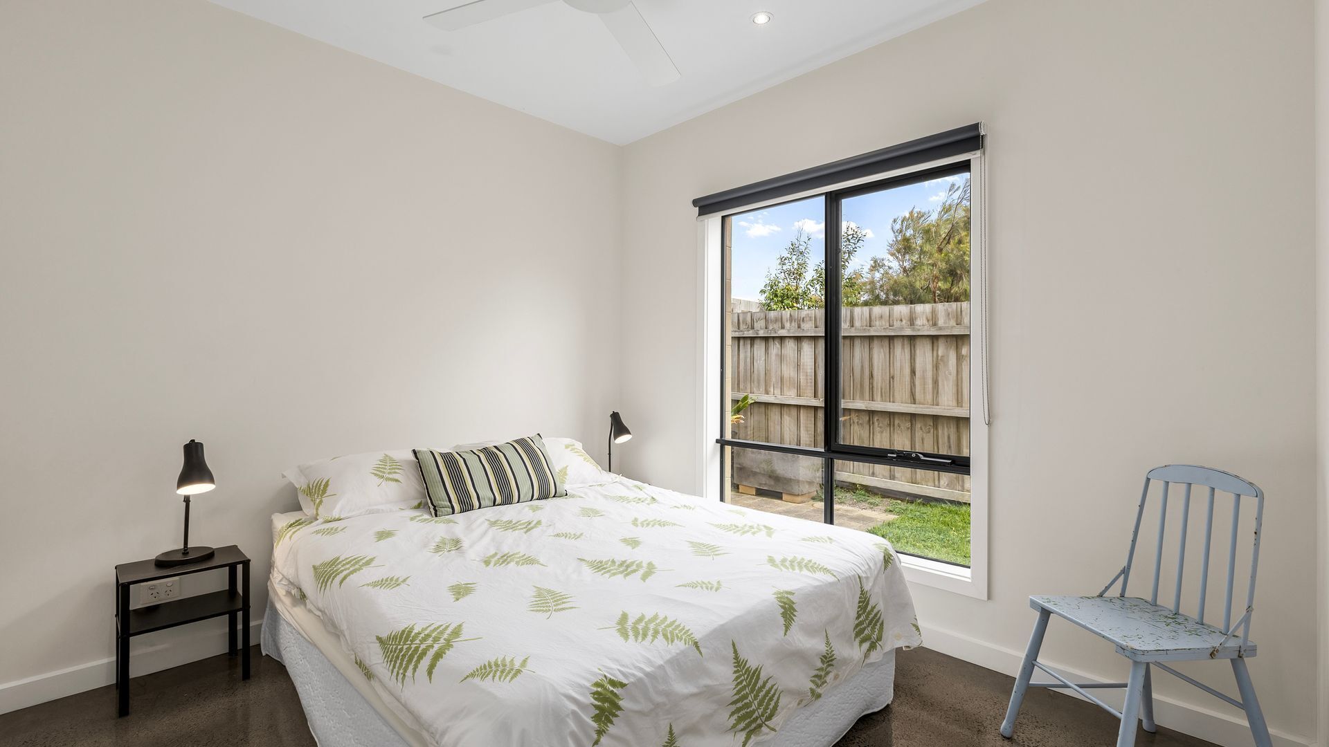 A bedroom with a bed, a nightstand, a chair, and a large window with a view of a wooden fence and greenery.