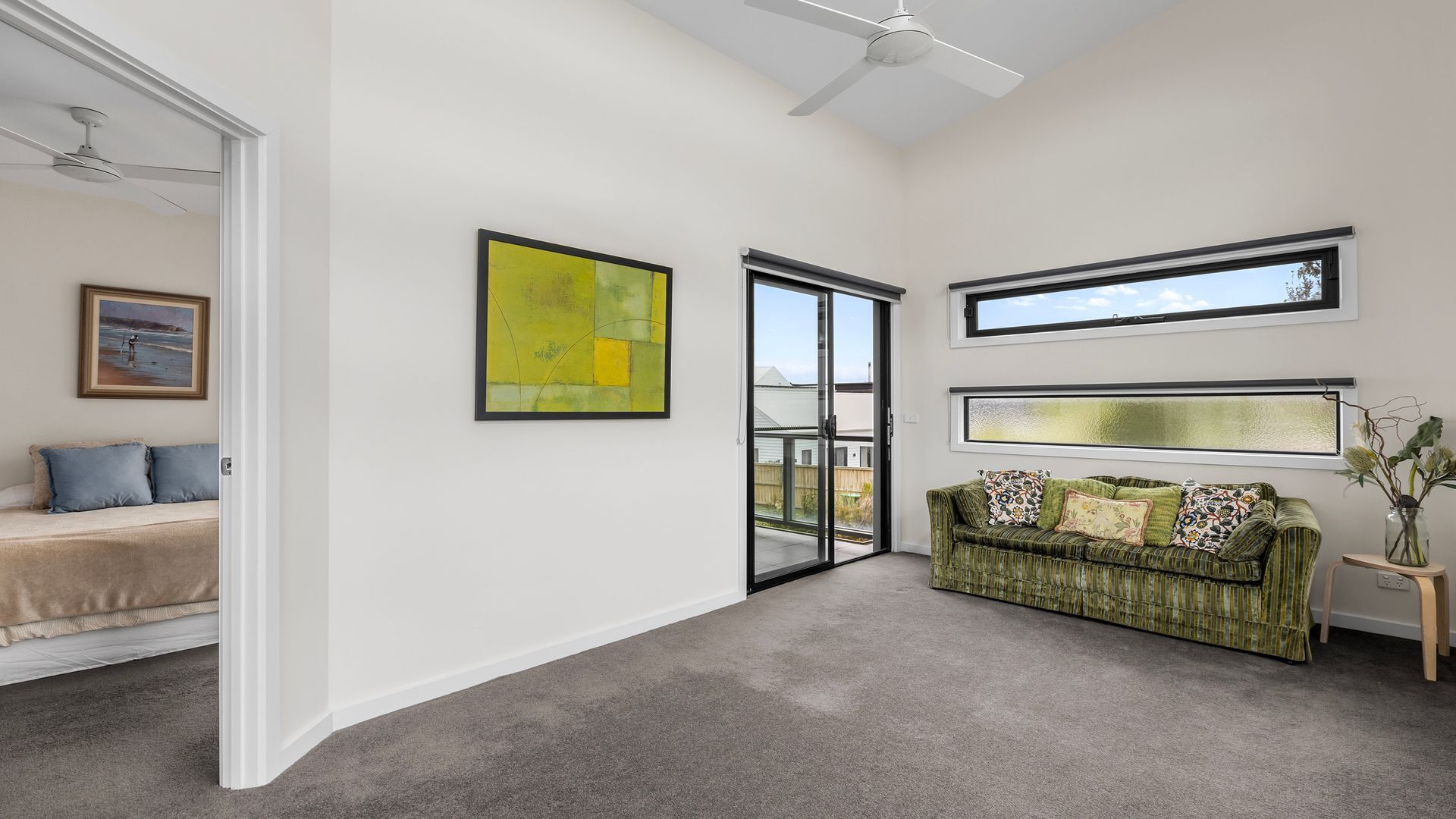 A spacious room with a green sofa, a framed abstract painting, and a large window offering a view of the outdoors.