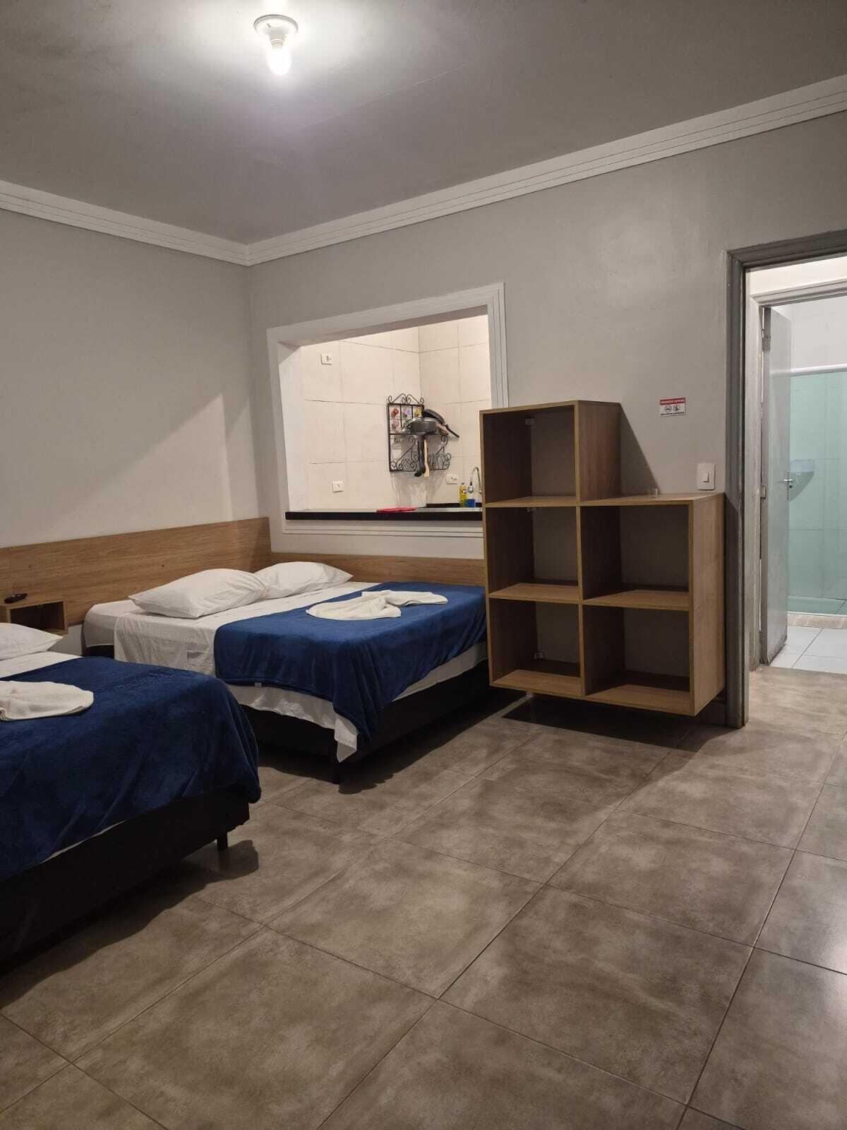 Quarto de hotel com duas camas, uma kitchenette pequena, uma estante de madeira e uma porta de banheiro.