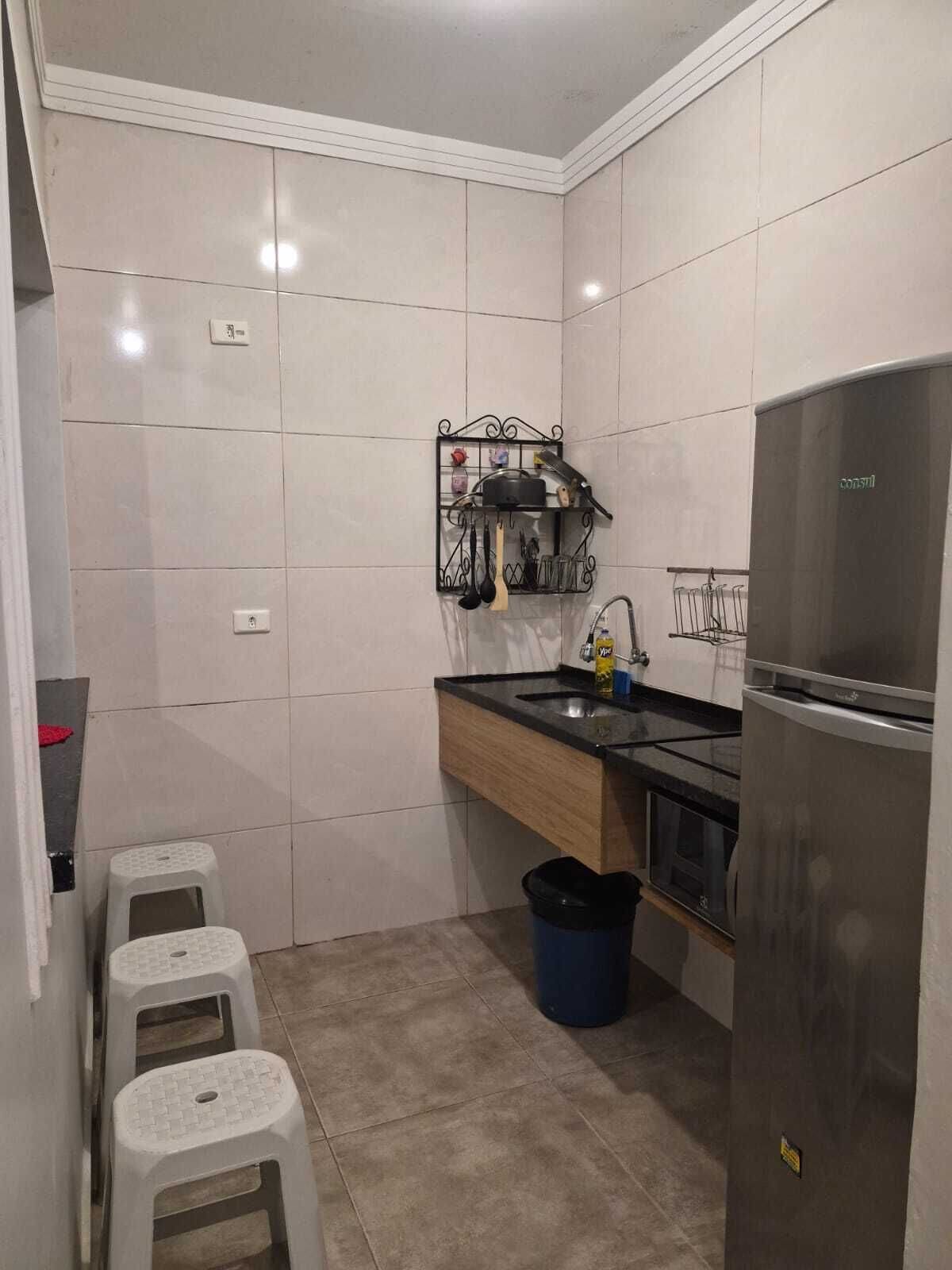 A compact kitchen with a stainless steel refrigerator, a black countertop, and a metal rack for utensils. There are three white stools and a blue trash bin.