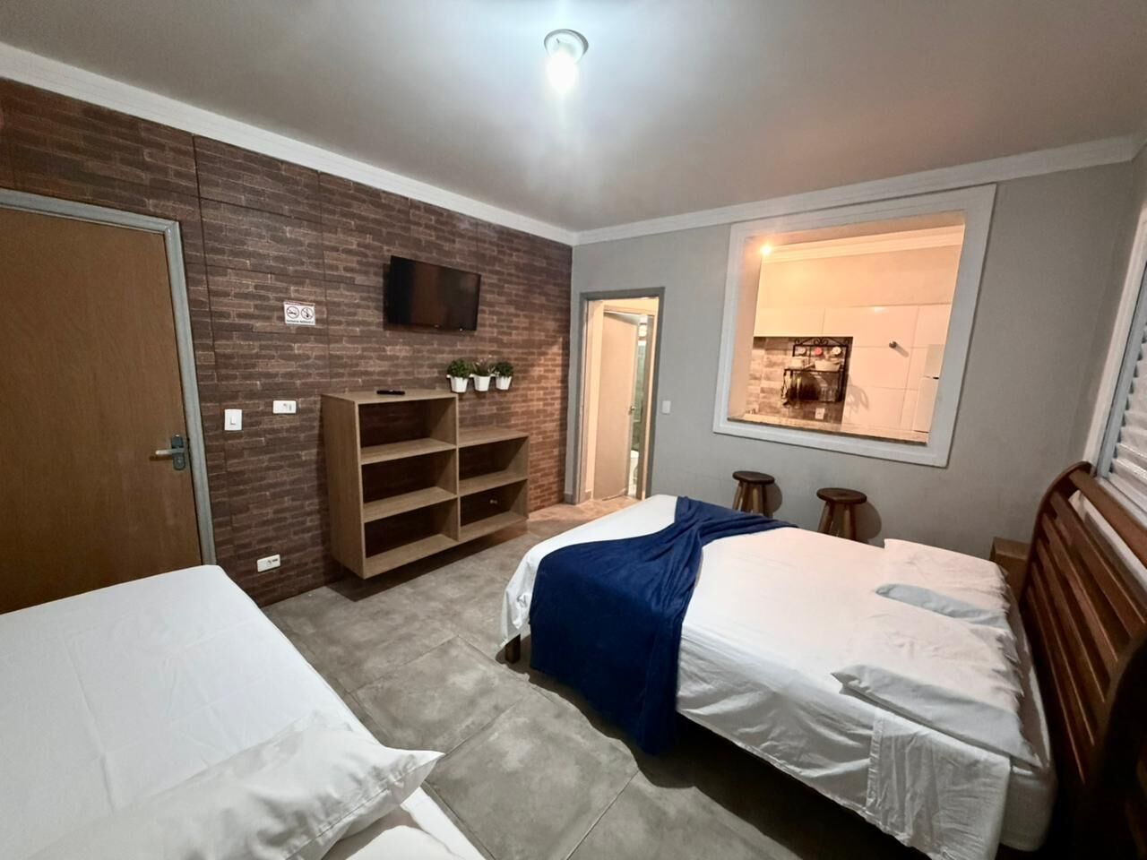 Un dormitorio con una cama, un armario de madera, un televisor montado en la pared y una ventana con vista a una cocina.