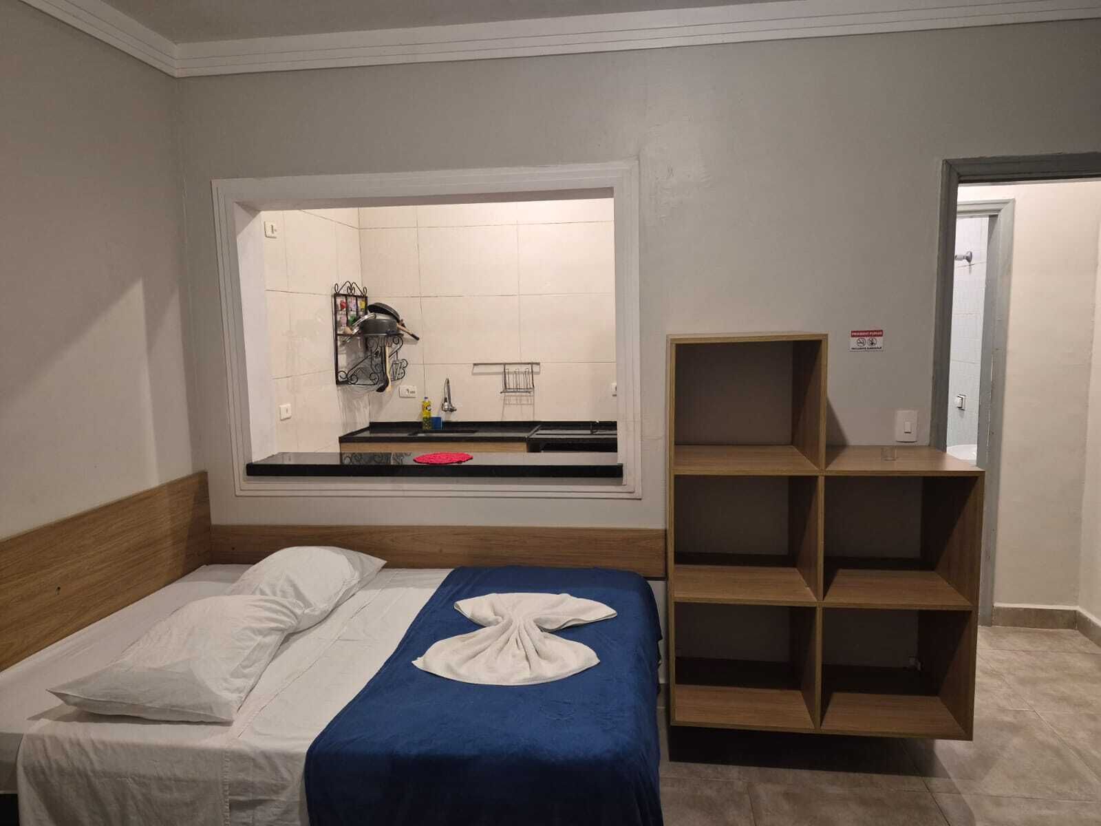 A hotel room with a bed, a small kitchenette, a wooden shelf unit, and a view of the bathroom.