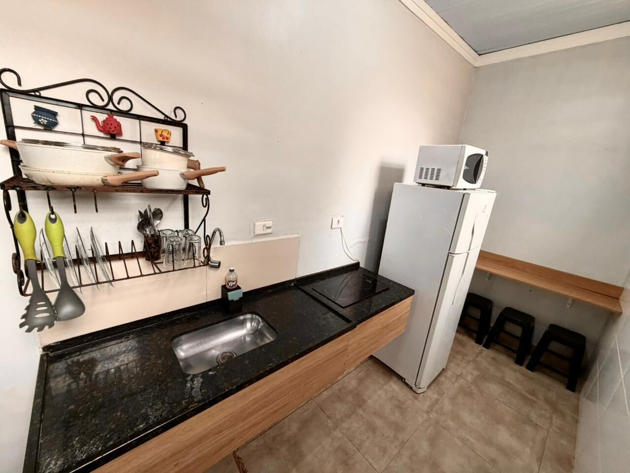 A compact kitchen with a black countertop, a sink, a white refrigerator, and a shelf with kitchen utensils.