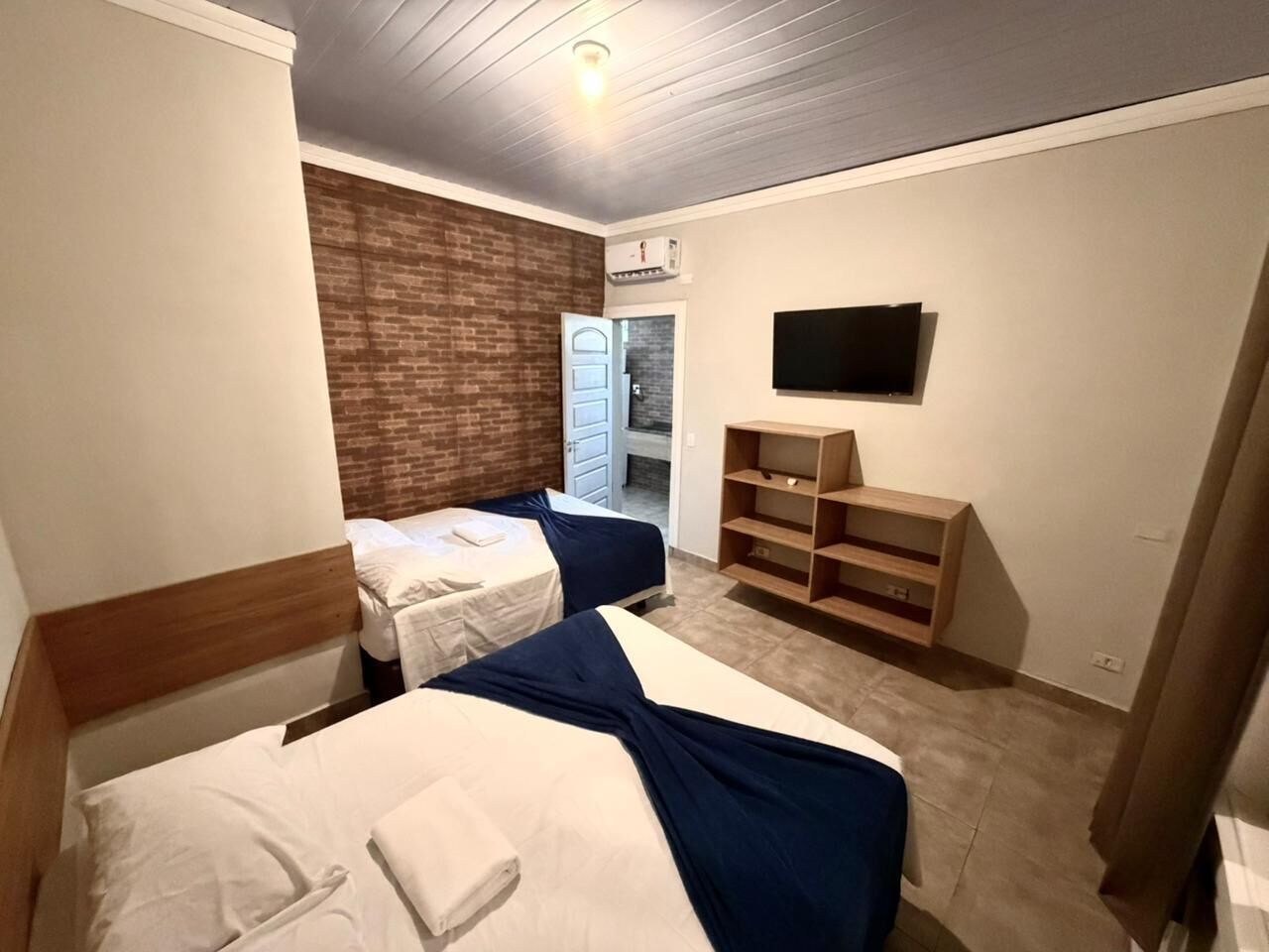 A hotel room with two beds, a brick accent wall, a TV, and a wooden shelf.
