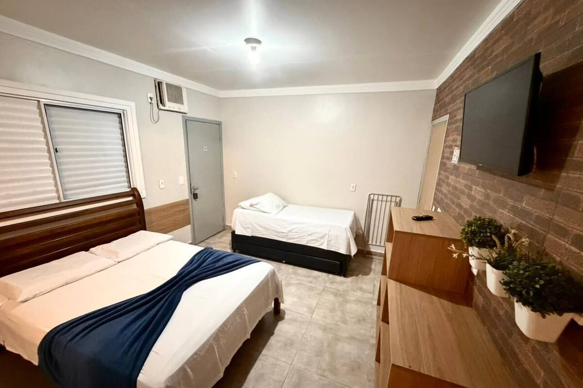 Une chambre avec deux lits, une commode en bois, un téléviseur fixé au mur et une petite fenêtre.