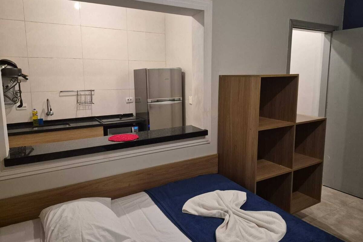 Une petite pièce comprenant un coin cuisine, un lit avec une couette bleue et blanche, et une étagère en bois.