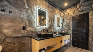A bathroom with a rustic stone wall, two sinks with black countertops, wooden vanity, and a black door.