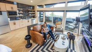 A modern living room with a sofa, a coffee table, and a TV. There is a kitchen area with cabinets and a microwave.
