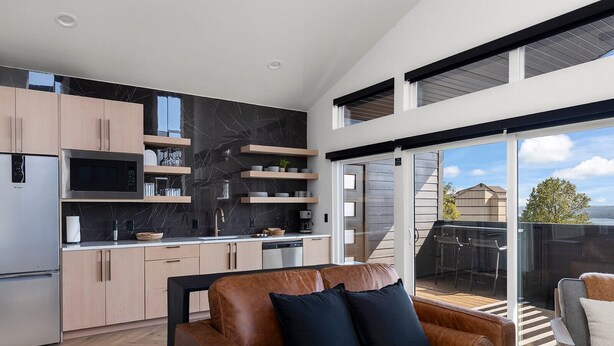 A modern kitchen with a black marble backsplash, stainless steel appliances, and wooden cabinets. There is a brown leather sofa with black cushions in the living area, and a sliding glass door leading to a balcony with a view of the sky and a tree. - Branson's Perfect Luxury Romantic Getaway (Branson)