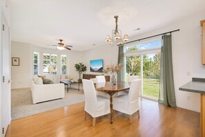 A spacious living room with a wooden dining table, white chairs, a ceiling fan, and a large window with a view of the outdoors.