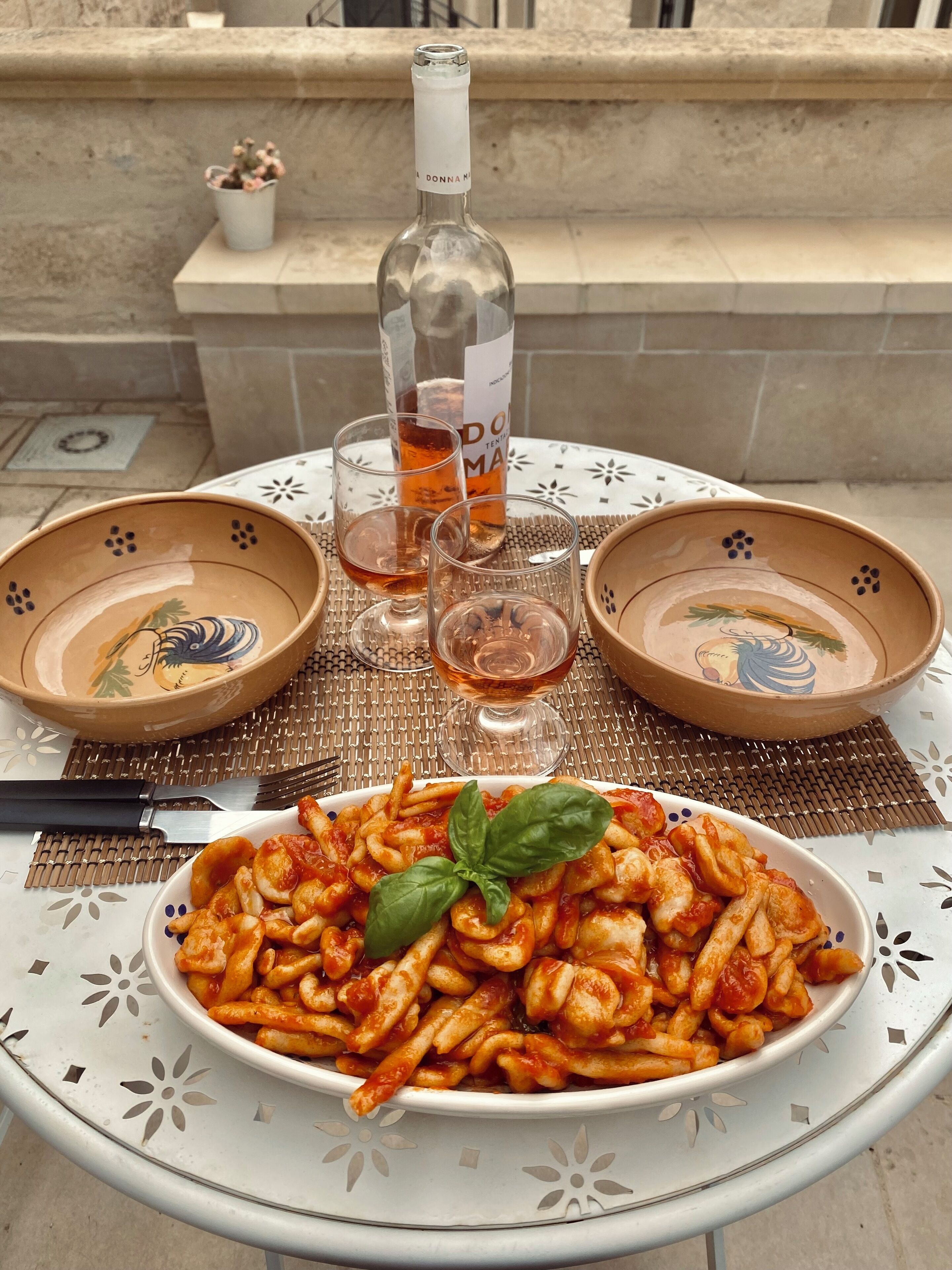 Una mesa con un plato de pasta, dos vasos, una botella de vino y dos cuencos decorativos.