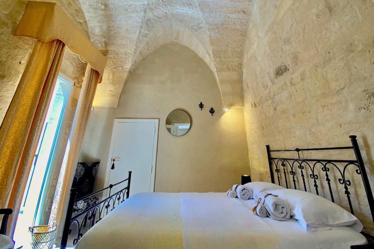 A room with a high arched ceiling, a bed with white linens, a round mirror on the wall, and a door.