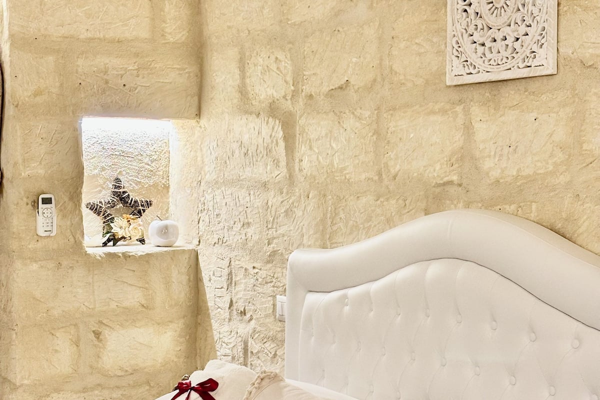 A bedroom with a stone wall, a white bed with tufted headboard, and a decorative wall piece.
