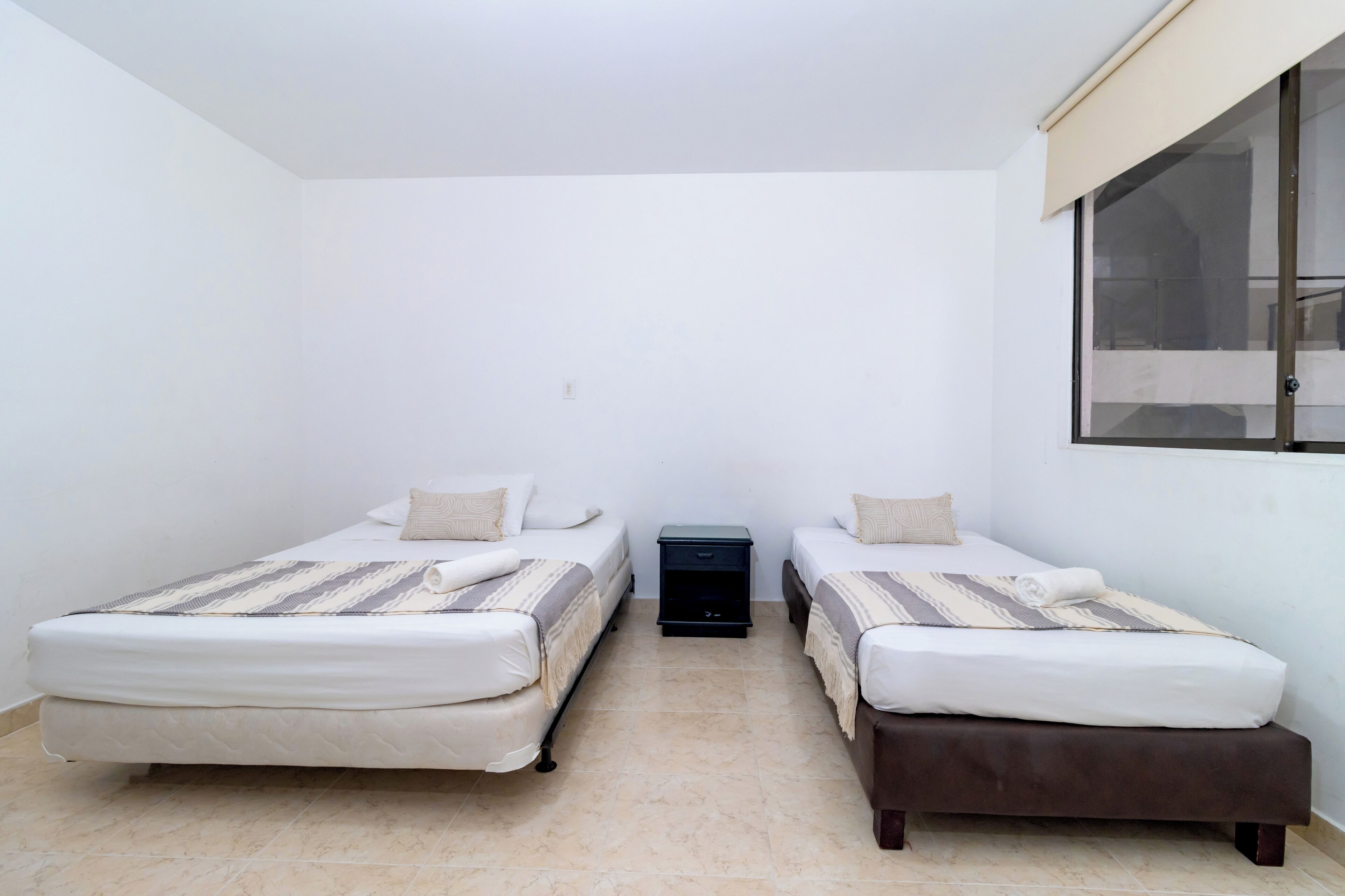 Une petite chambre propre, avec deux lits simples, une table de chevet et une fenêtre.