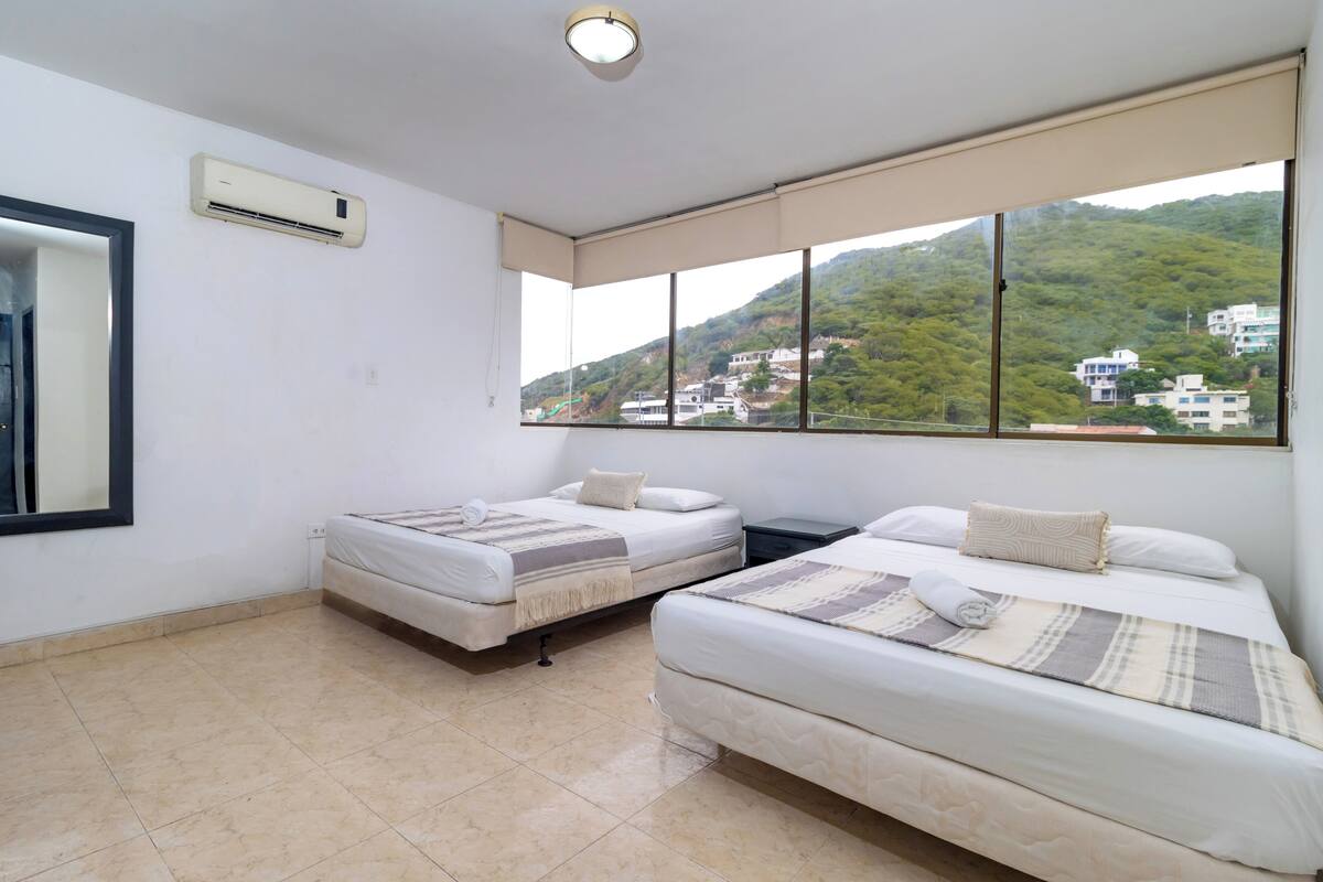 A hotel room with two beds, a large window with a view of a hillside, and an air conditioning unit on the wall.