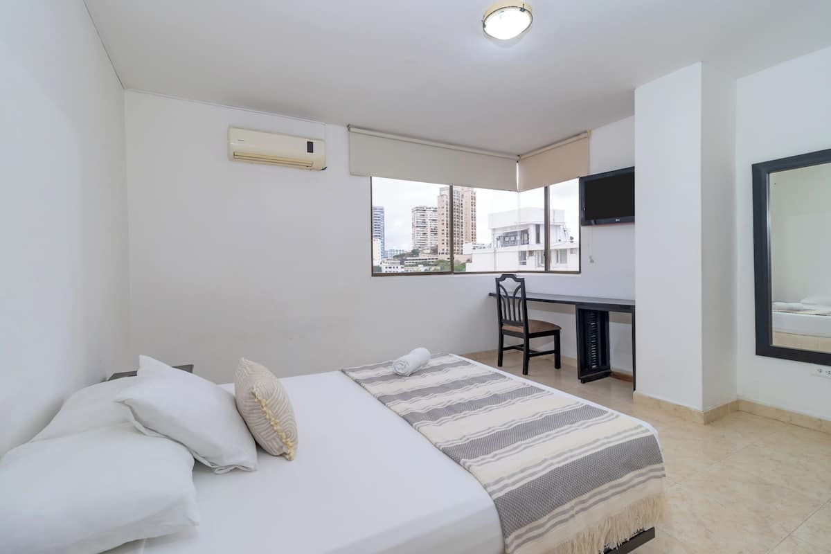A hotel room with a bed, a desk, a chair, a TV, and a window with a city view.
