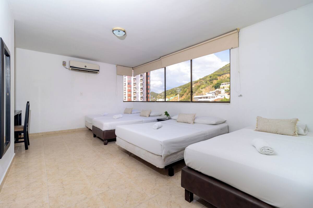A hotel room with three beds, a window with a view of buildings and hills, an air conditioning unit, and a small table with a chair.
