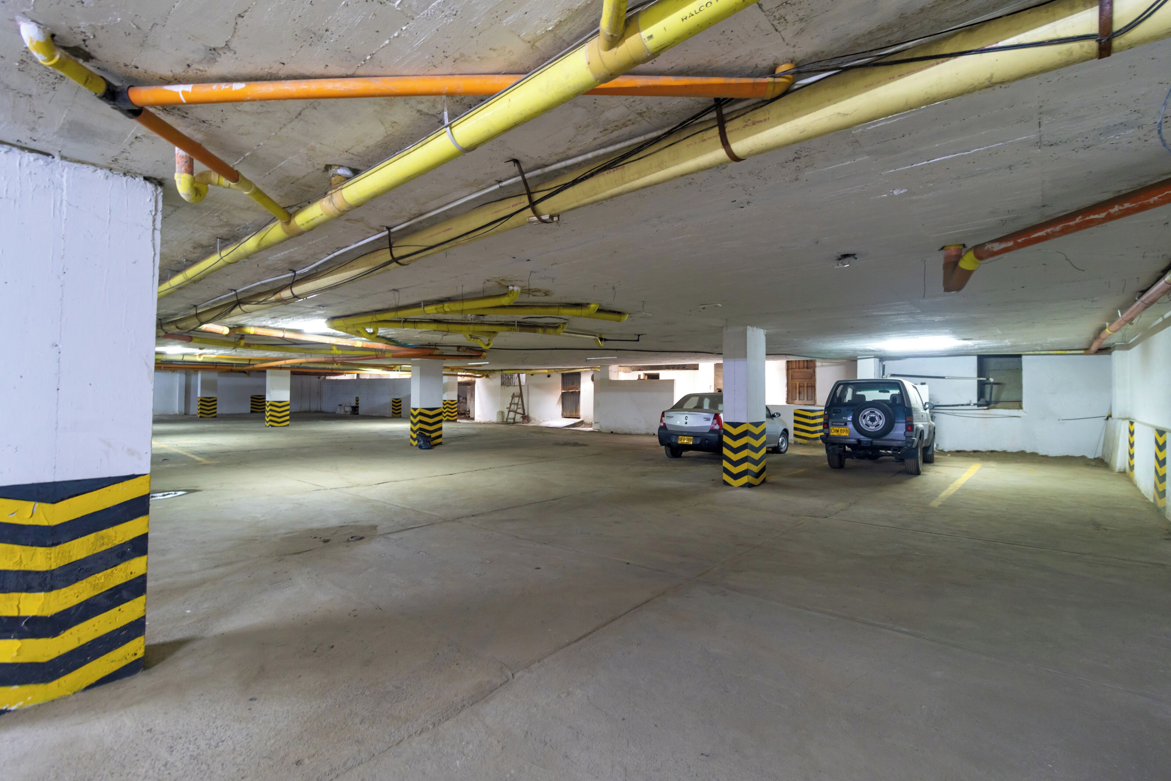 Un parking souterrain avec des piliers en béton, des marquages de sécurité jaunes et noirs en bandes, et plusieurs voitures garées.