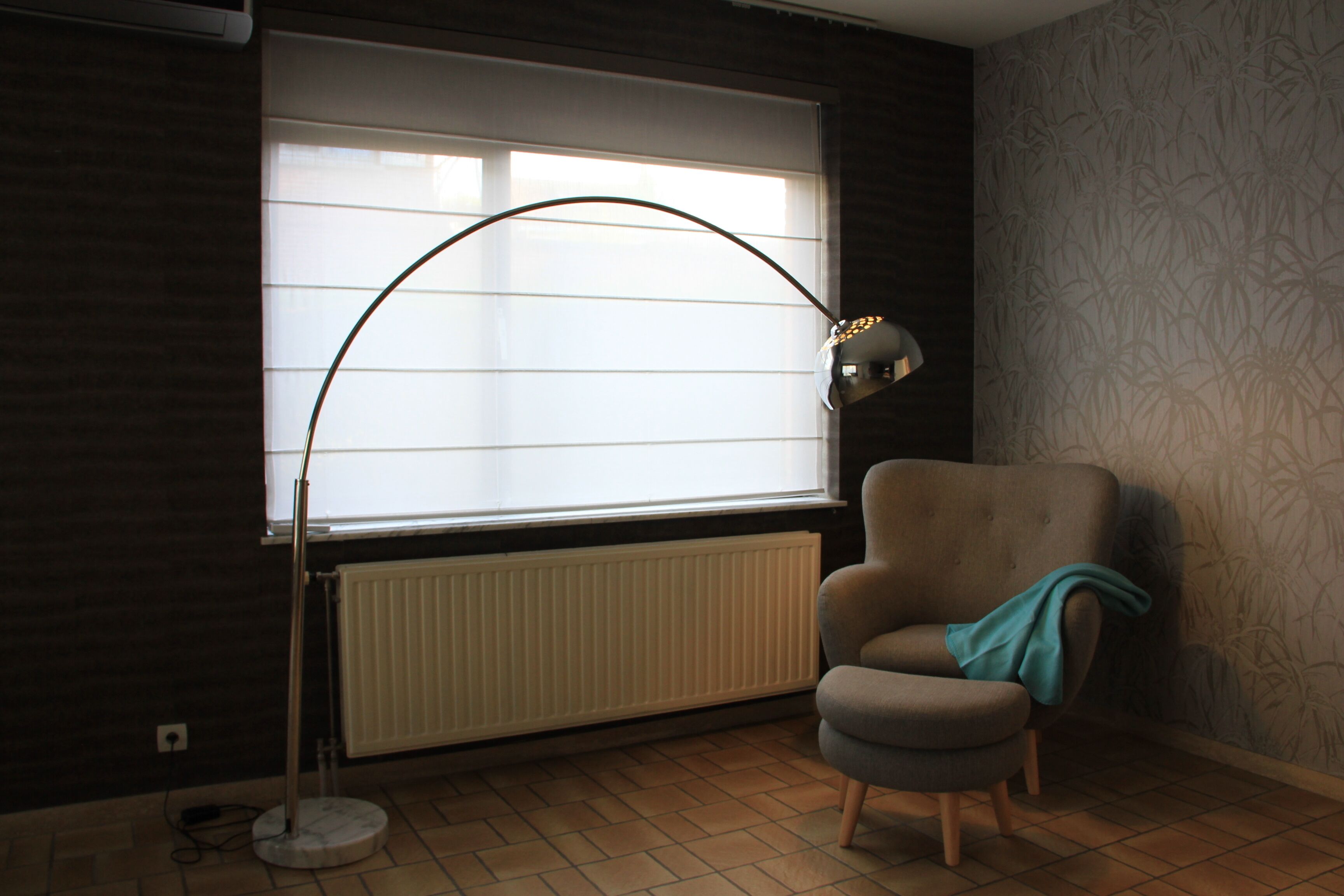 A room with a radiator, a window with blinds, a chair, and a floor lamp.