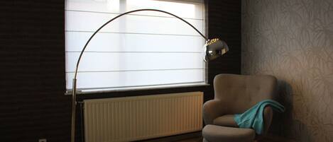 A room with a radiator, a window with blinds, a chair, and a floor lamp.