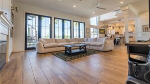 Ein geräumiger Wohnbereich mit einem großen Ecksofa, einem Couchtisch und einem Esstisch. Deckenventilatoren, große Fenster und ein Blick ins Freie sind ebenfalls vorhanden.