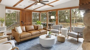 A sunroom with a wooden ceiling, large windows, a brown leather sofa, a white coffee table, and a gray armchair.