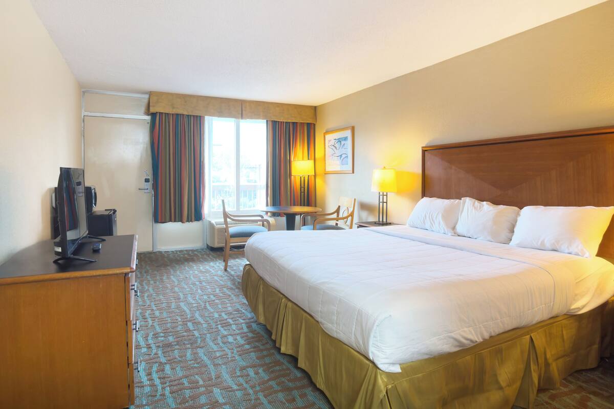 Ein Hotelzimmer mit einem großen Bett, einem Schreibtisch mit Computer, einem Stuhl, einem Tisch und einem Fenster mit Vorhängen.