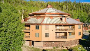 A multi-story building with a sloped roof, balconies, and a stone foundation, surrounded by dense forest. - Timbernest 1A - Cozy & Convenient Mountain Condo (Brian Head)
