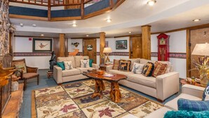 A spacious living room with a high ceiling, wooden beams, and a staircase. There are two sofas, a coffee table, and a rug with a floral pattern. - Timbernest 1A - Cozy & Convenient Mountain Condo (Brian Head)