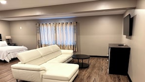 A modern hotel room with a beige sofa, a small round table, a black speaker, and a bed with white bedding. - Four Seasons Inn (Gallipolis)
