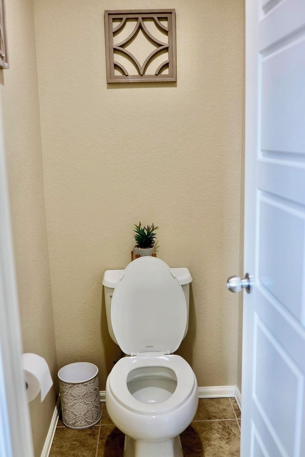 A small bathroom with a toilet, a decorative wall panel, a potted plant, and a trash can.