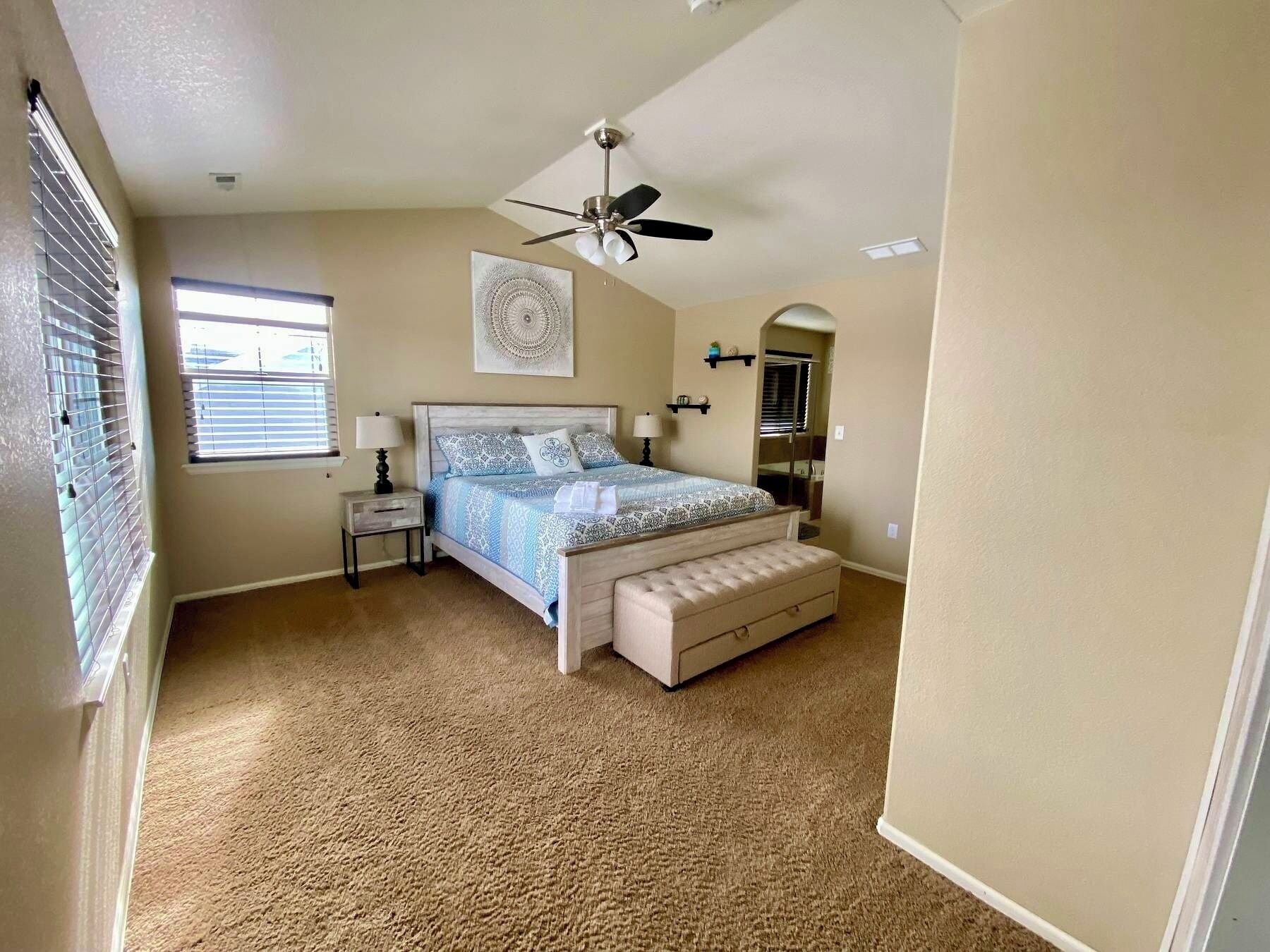 A bedroom with a bed, bedside tables, a ceiling fan, a window with blinds, a wall-mounted mirror, and a carpeted floor.