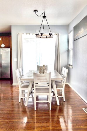 Un espace repas avec une table et des chaises en bois, un lustre et une vue sur l’extérieur grâce à de grandes fenêtres.