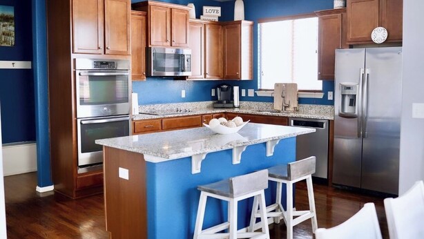 A modern kitchen with wooden cabinets, stainless steel appliances, a granite countertop, and a blue accent wall. - Immaculate Home in Aurora (Aurora)