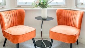 Une pièce comprenant deux fauteuils recouverts d’un tissu orange, une petite table ronde sur laquelle est posé un vase blanc rempli de fleurs, et une grande fenêtre à carreaux blancs ornée d’une grille décorative.